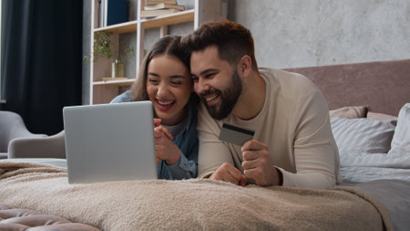 Caucasian happy couple woman and man relax on bed with laptop shopping online use bank credit card pay order buying in internet booking financial service e-commerce app winning success spending moneyの写真素材