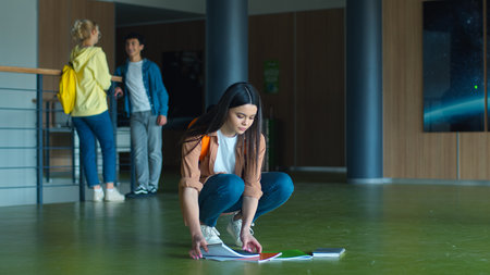 Caucasian woman girl female schoolgirl student dropped books fell on floor in school academy corridor pick up copybooks textbooks friends boy guy classmates talking on background educationの写真素材