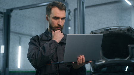 Serious focused Caucasian man guy technician auto mechanic engineer check repairing car problem computer diagnostics electric automobile engine with laptop checking maintenance in garage vehicle shopの写真素材
