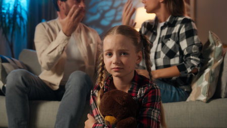 Frustrated stressed upset Caucasian daughter child kid girl holding teddy bear parents quarrel conflict shouting at home evening night domestic family man father arguing mother womanの写真素材