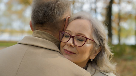 Gray-haired 60s mature Caucasian woman wife embrace cuddle beloved man husband in park autumn nature elderly couple hugs outdoors satisfied happy female embracing male tender love support at old ageの写真素材