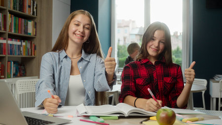 Smiling Caucasian girls teens women schoolgirls students pupils high school learning studying together at desk university college writing lesson looking at camera thumbs up like approve good educationの写真素材