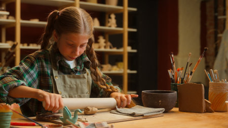 Young creative Caucasian little girl teen child junior kid schoolgirl student potter artist kneading clay material use rolling pin creating pottery artwork handmade ceramic art lesson inside workshopの写真素材