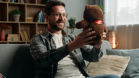 Happy smiling Caucasian man male guy father dad at home couch holding cute toy Teddy bear preparing gift present childish anticipate prepare to child children baby custody adoption donation volunteerの写真素材
