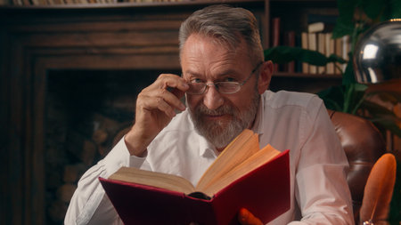 Satisfied old Caucasian man holding book textbook reading reader literature looking at camera smiling male home library pensioner grandfather elderly senior education study businessman business indoorの写真素材