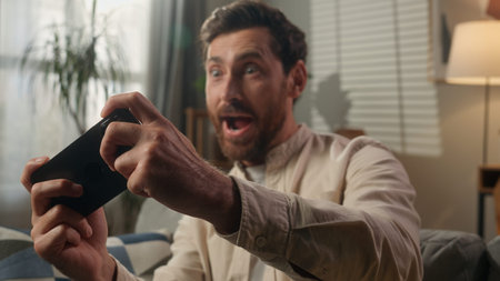 Excited happy Caucasian adult man gamer playing mobile phone on couch sofa living room smiling guy...