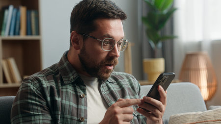 Portrait adult looking at mobile phone shock reaction amazed bad news close mouth with hand problem online indoors. Closeup male in apartment holding using smartphone shocked reaction at home scaredの写真素材