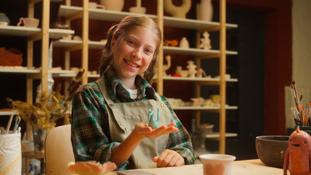 Joyful cheerful smiling Caucasian little girl artist sculptor ceramist child school kid student daughter holding hands ceramic swan bird clay creation artwork pottery art looking at camera in workshopの写真素材