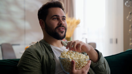 Happy relaxed Caucasian bearded model business man male guy businessman homeowner client sitting sofa at home eating snack popcorn bowl watching TV television series using remote controller film movieの写真素材