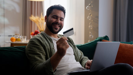 Happy smiling Caucasian man male guy shopper client customer homeowner using laptop typing computer holding credit card bank paying online ecommerce shopping internet buy transaction indoor home roomの写真素材
