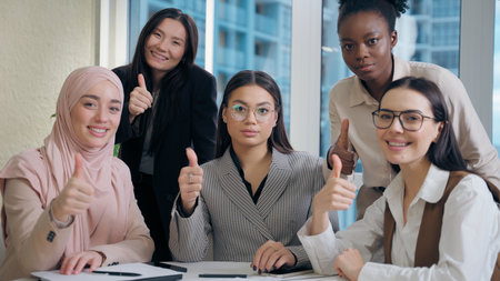 Brainstorming meeting multiethnic women females diversity team five girls coworkers businesswomen business colleagues discuss documents startup looking at camera showing thumb up gesture indoor officeの写真素材