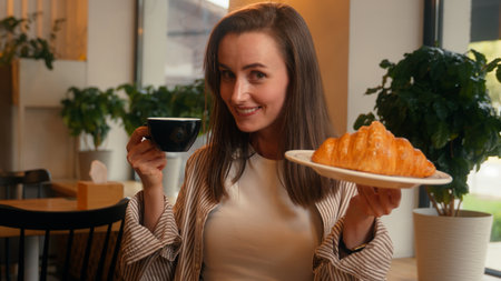 Happy smiling positive Caucasian woman female cafe visitor girl in cafeteria breakfast lunch holding cup of tea coffee aromatic drink and plate with tasty croissant looking at camera calories foodの写真素材