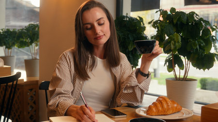 Caucasian woman in cafe inspired writing notes in notebook drinking coffee lunch happy female girl at cafeteria table drink tea with croissant write diary information noting idea handwriting planの写真素材