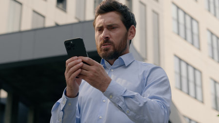 Mad unhappy middle-aged mature businessman man business male feeling annoyed using smartphone in city angry employer problem mobile phone spam message irritated frustrated by bad signal outdoors workの写真素材