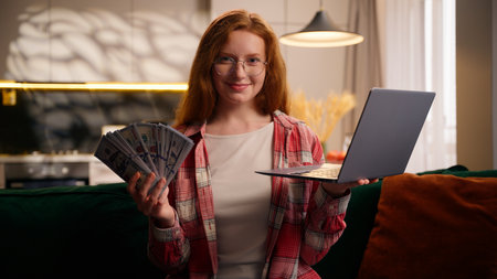 Portrait smiling happy Caucasian woman looking at camera celebrate holding money cash prize bet winning reward with laptop computer girl female user gamer gambler achieve with dollars casino at homeの写真素材