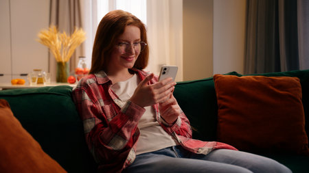 Positive happy smiling laughing Caucasian woman female girl user gadget addict using online app smartphone social media chat communication sitting on home couch typing message mobile phone cheerfulの写真素材