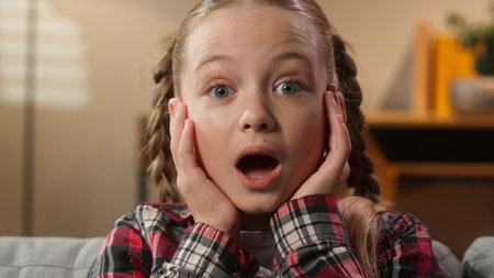 Little Caucasian girl child kid looking at camera with big eyes saying wow amazing news reaction portrait daughter in living room home look holding head shock wonder surprise unbelievable astonishの写真素材