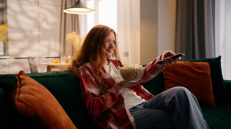 Caucasian girl female woman happy falling on couch cozy home sitting on sofa rest chilling at living room eating popcorn watching TV film movie television relaxing weekend apartment insurance mortgageの写真素材