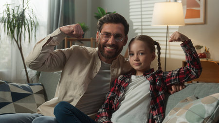 Happy Caucasian family funny smiling adult strong father with cute daughter dad man kid girl child at home at couch looking at camera showing hands biceps champions gesture health muscles sport fitの写真素材