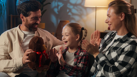 Happy Caucasian family on sofa in evening home father giving teddy bear to daughter kid cheerful moment parents applauding smiling child girl hugging toy love celebration birthday gift childcareの写真素材