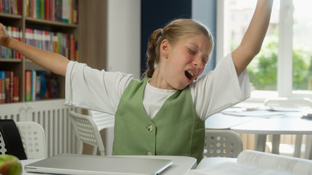 Tired girl in school child kid exhausted bored schoolgirl learner pupil lazy sleepy Caucasian student yawning stretching hands sleep lack energy in classroom boring homework exam indoors educationの写真素材