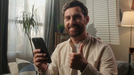 Caucasian man satisfied mobile phone using modern app male guy smiling businessman homeowner relax on couch browsing smartphone device thumb up good recommend order food delivery online shopping homeの写真素材