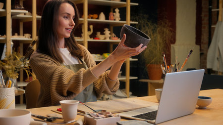 Potter Caucasian girl woman artist sculptor saleswoman small business owner ceramics sale laptop chatting online shopping handmade crockery craft bowl commerce seller typing computer in art workshopの写真素材