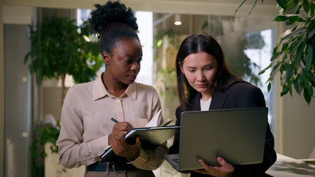 African Asian business women girls colleagues multiracial businesswomen in office teamwork laptop project writing tasks notebook Chinese Korean American co-workers two female managers corporate workの写真素材