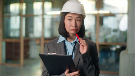 Thoughtful woman Asian Chinese Japanese girl builder architect engineer thinking Korean businesswoman technician contractor female in hard hat pensive writing notes building plan document constructionの写真素材