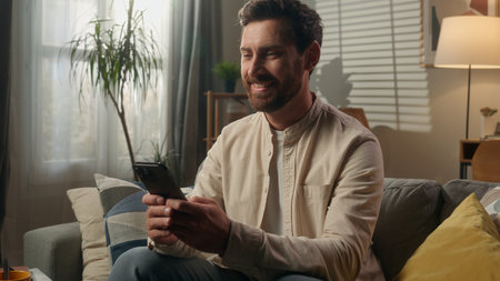 Smiling man gadget addict businessman Caucasian guy homeowner relax using smartphone on couch living room happy male browsing mobile phone online shopping social media laughing enjoy message positiveの写真素材