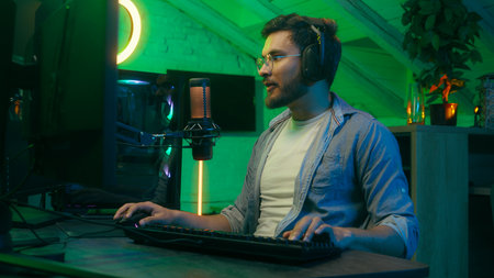 Caucasian guy male man streamer professional gamer wearing headphones playing virtual video game online play gaming sitting at computer monitor at night home neon technology streaming enjoy hobbyの写真素材