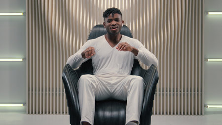 African American guy man ethnic male putting wireless headphones in ears listening music dancing waving arms moving hands dance to audio song in earphones sitting in futuristic white studio futurismの写真素材