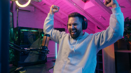 Happy young man Caucasian guy male celebrating victory gaming on computer stream with headphones and microphone in neon room joyful gamer excited after winning online battle rejoice reaction winnerの写真素材