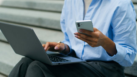 Unrecognizable African American student girl female business woman businesswoman lady sitting in city urban outdoors work online typing laptop pc computer using mobile phone smartphone multitaskingの写真素材