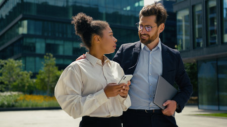Two business people in city African businesswoman using phone walk Caucasian businessman man talking to female smiling put hand on shoulder woman communicationの写真素材