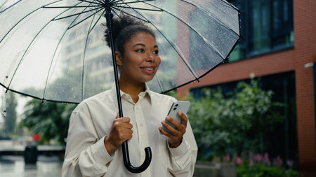 African American woman texting mobile phone smartphone chat online message looking around waiting taxi order meeting female girl businesswoman city outdoors smiling business entrepreneur rain umbrellaの写真素材