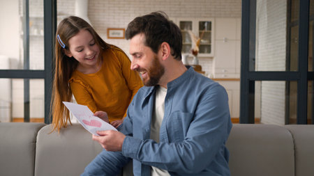 Carefree glad Caucasian daughter kid little girl running to father dad adult man surprise hug. Parent male receiving Fathers Day card present from generous child smile family couch at home love trustの写真素材