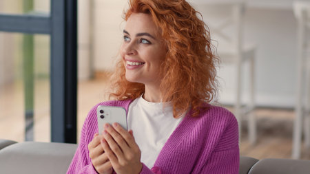 Happy smiling carefree Caucasian woman lady girl indoors at home couch sofa female using mobile phone smartphone browsing app social media network smile looking away thinking ponder shopping onlineの写真素材