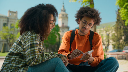 African American ethnic students people classmates woman girl lady female man male guy sit outside in city talking discuss mobile phone looking pensive smartphone use university college high schoolの写真素材