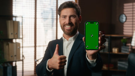 Smiling happy middle aged business man showing green screen mobile phone like gesture thumb up holding smartphone chroma key display success achieve mature businessman male looking at camera in officeの写真素材