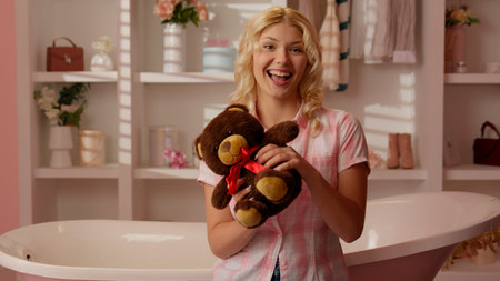 Smiling joyful Caucasian girl female teenager woman in pink candy glamour bathroom looking at camera holding Teddy Bear playing showing fluffy toy happy carefree indoors present gift valentines dayの写真素材