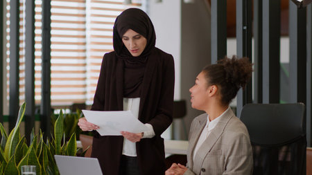 Arabian Muslim in hijab and African American women ladies business girls businesswomen in office discuss papers corporate documents female teamwork boss coach mentor help teach intern employee consultの写真素材