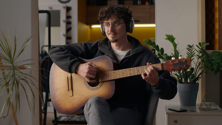 Smiling Caucasian musician guy playing guitar solo at home hipster man in headphones male hobby...