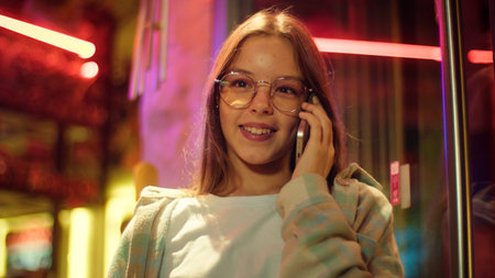 Glad teenage young woman talking mobile phone in city neon female student smile smartphone answer...