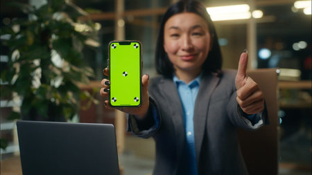 Asian business woman businesswoman holding mobile phone showing smartphone green screen chroma key display female girl smiling thumb up advertise app recommend office workplaceの写真素材