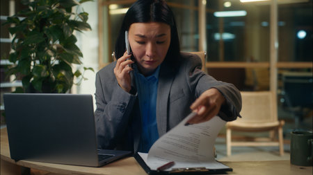 Worried Asian Korean Chinese Japanese woman talking mobile phone smartphone call girl businesswoman business problem searching information documents paperwork female in office manager employer troubleの写真素材