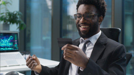 Happy joyful African American man holding banking credit card online buying consumer payment computer buyer businessman male guy shopper business manager employee entrepreneur office client purchaseの写真素材
