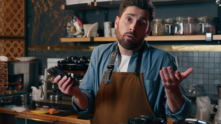 Confused Caucasian barista man holding mobile phone shrug shoulders in cafeteria guy waiter unsure...