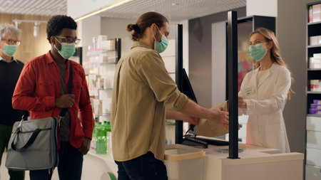 Pharmacy cashier counter woman pharmacist in mask covid pandemic help customers buying pills health care medication in drugstore shop medical purchase paying contactless to terminal medicine serviceの写真素材