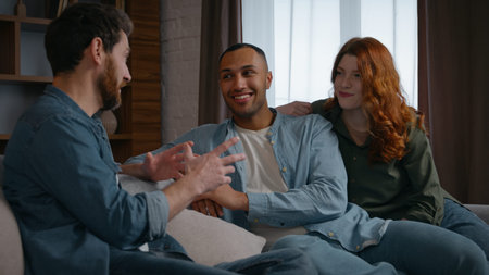 Three multiracial friends at home party talking Caucasian man sharing news Latino African American...